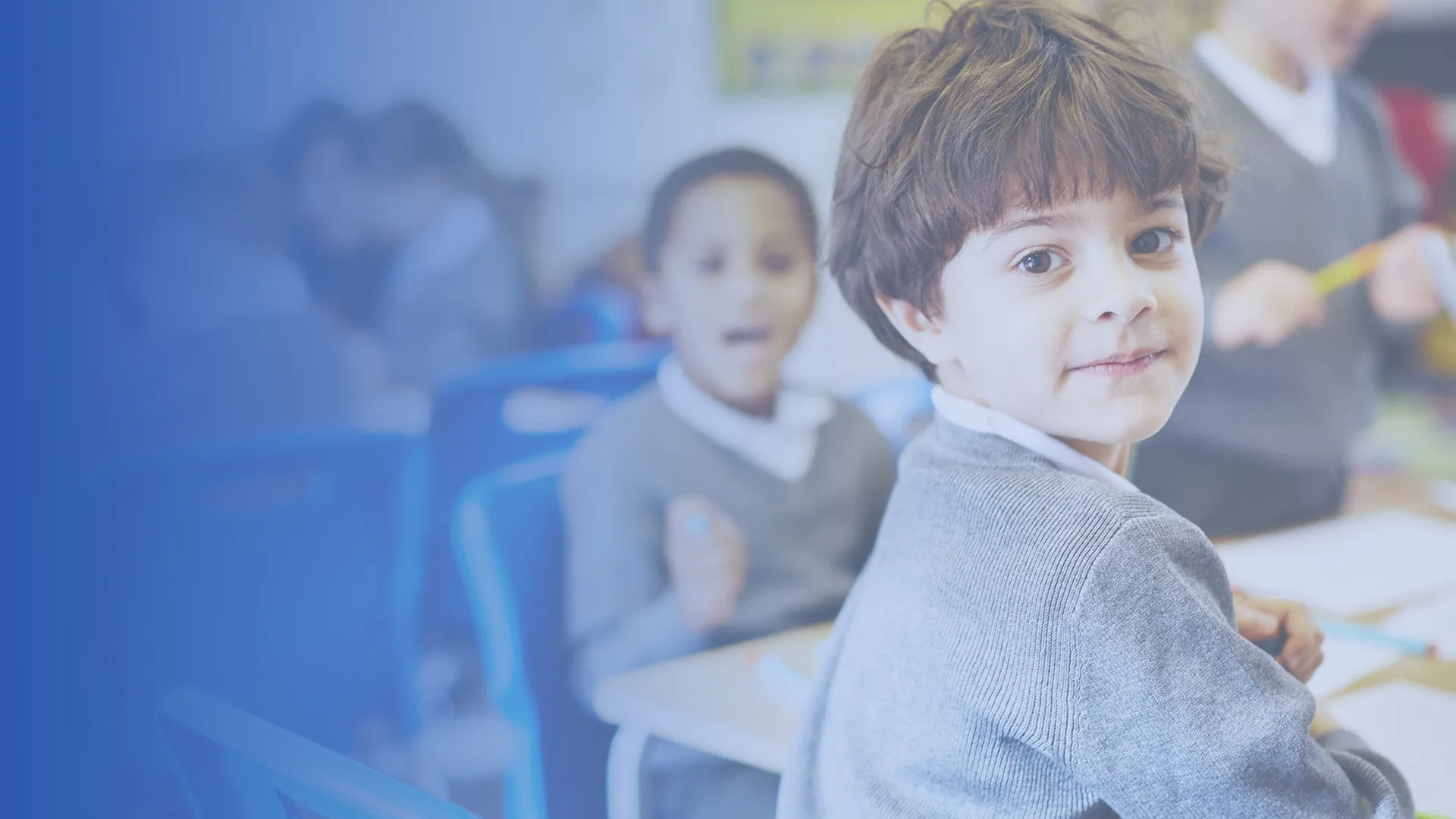 A smiling boy in school uniform looking over his shoulder in a primary classroom.