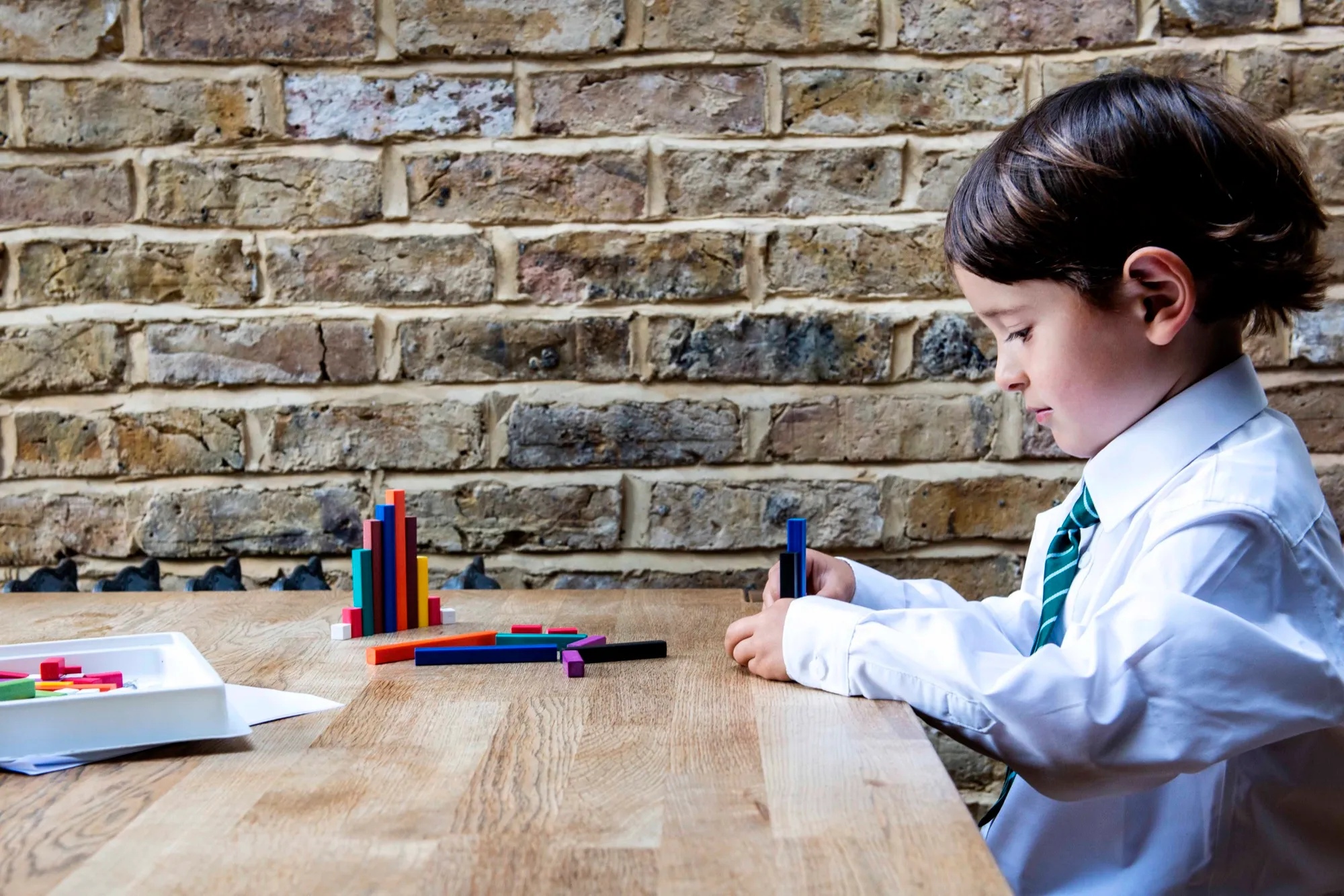 A boy in school uniform sitting at a wooden table, working with coloured counting rods.
