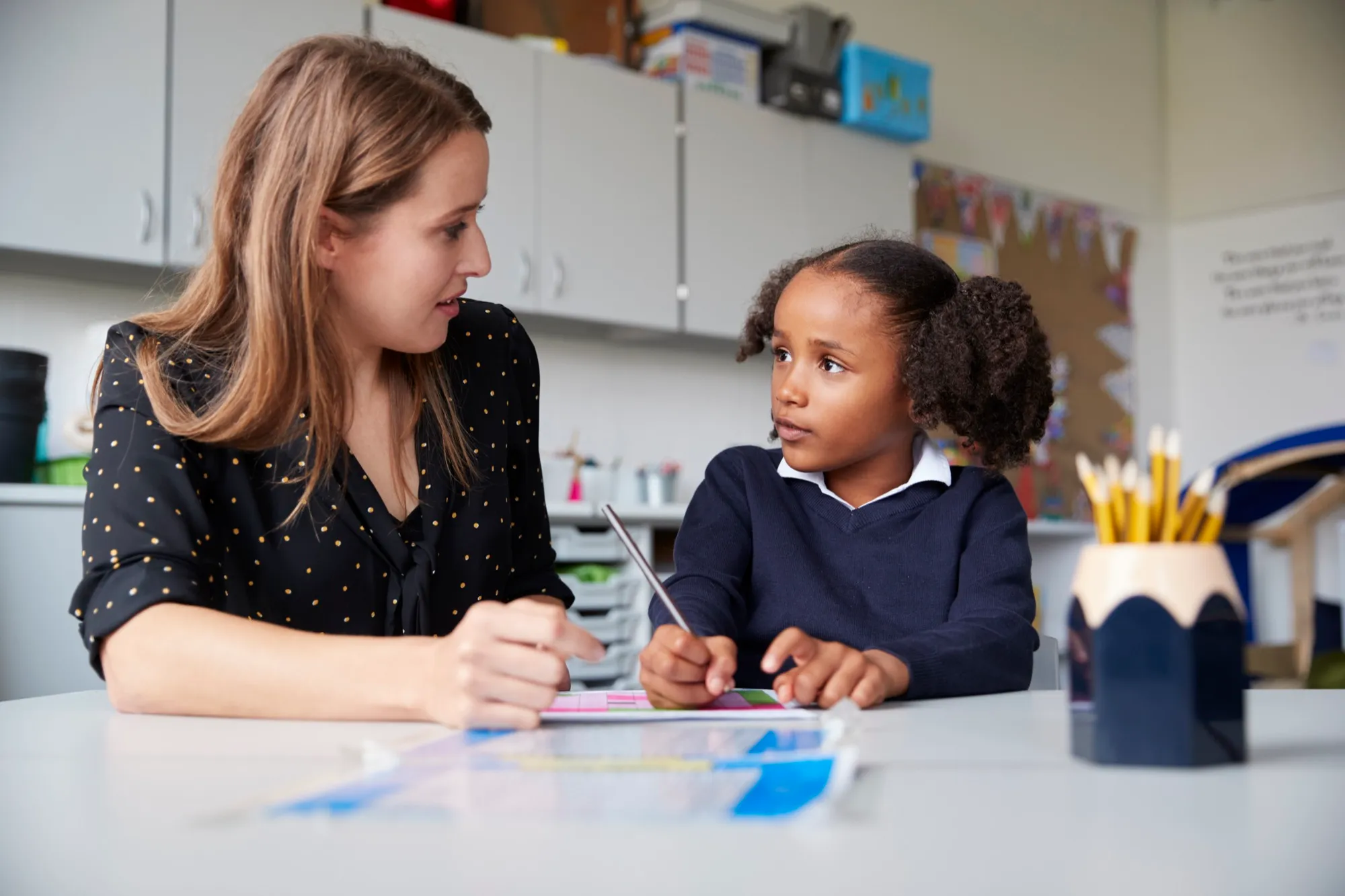 A teacher and a girl in school uniform sitting side by side at a table, working through a page together.